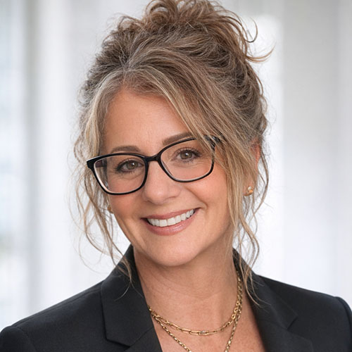 A woman with curly, shoulder-length hair wearing glasses and a black blazer, smiling warmly in a well-lit indoor setting.