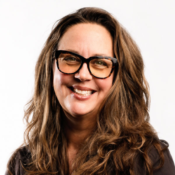 Smiling woman with long, wavy brown hair and glasses, wearing a dark shirt, against a white background.