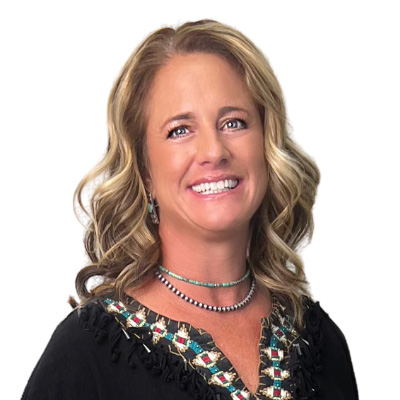 A smiling woman with wavy blonde hair is wearing a black embroidered top and layered necklaces. She is posed against a plain white background.