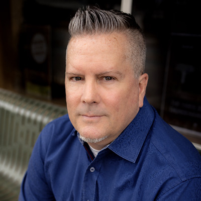 Close-up professional headshot of Kelly Messick, a man with short graying hair styled up and a slight soul patch, wearing a blue collared shirt.