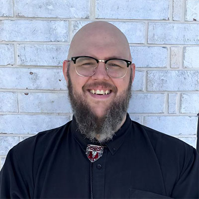 A headshot portrait of a smiling, bald man with a full graying beard, wearing browline glasses and a black button-up shirt decorated with a bolo tie, posed in front of a light gray brick wall.